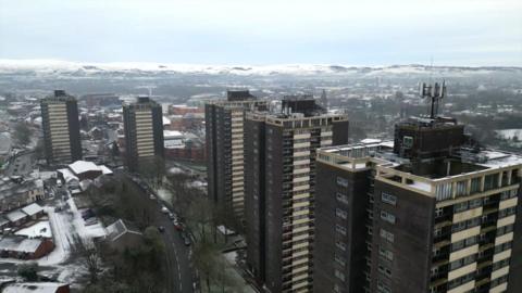 Six tower blocks can be seen to the right of the camera lining a road, with snowy hills in the far background