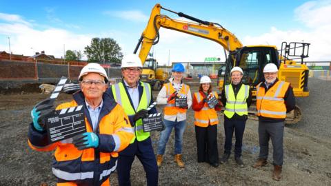 Six people wearing hi-vis jackets and hard hats on a building site. Four of them are holding black and white clapper boards, like those used on film sets at the start of filming. A yellow excavator is parked behind them.