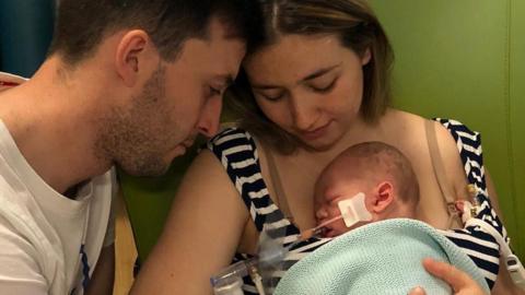 Kianty is holding Jacob wrapped in a blue blanket against her chest. The baby is attached to a breathing tube. Conor sits close to Kianty with his arm around hers which is holding the baby.