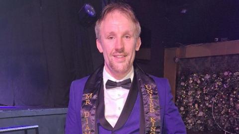A man wearing a purple tuxedo smiling at the camera. He is standing in front of stage and is wearing a sash that says Mr Diamond United Kingdom.