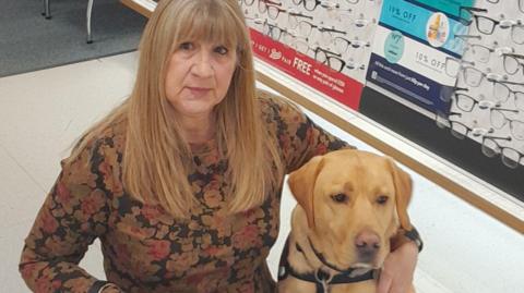 Linda on the left with mid-length blonde hair wearing a long-sleeve floral top. She has her arm around her hearing assistance dog Toffee who is a brown Labrador.