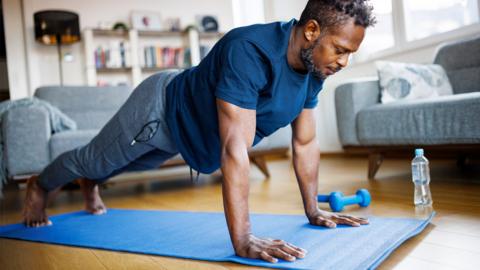 A man working out on an exercise mat in a plank position. He is wearing workout clothing and has a bottle of water and some weights beside him. 