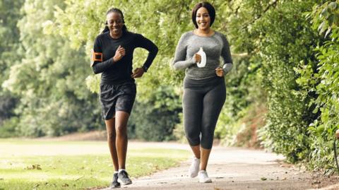 Two women wearing exercise gear, running on a path in a park, smiling 