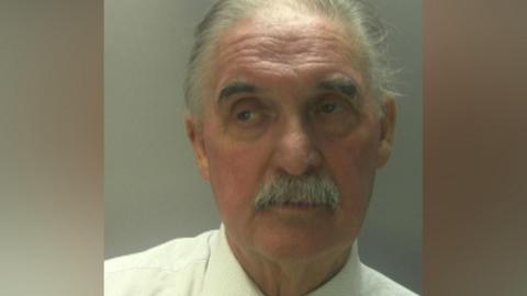 A man with grey hair and moustache. He is wearing a white shirt.