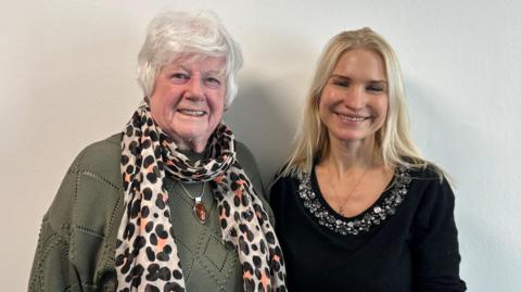 Two women - one with short grey hair and wearing a green jumper, a spotty scarf and a large amber necklace, and the other with long blonde hair and wearing a black jumper with a jewelled collar. They're standing next to each other and smiling at the camera against a white background.