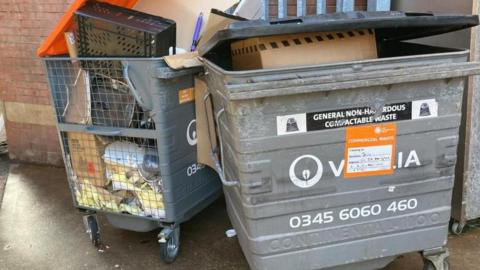 Two wheelie bins in the centre of Northampton, by a metal fence. A door is open to the right.