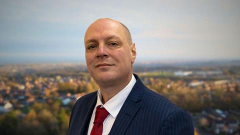 A bald man wearing a blue pin-stripped blazer, white shirt and red tie, standing in front of an aerial image of a blurred town