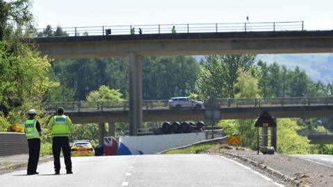 Two police officers stand in hi vis clothing bottom left of shot with an overturned lorry on the road in the distance below two concrete flyover bridges.
