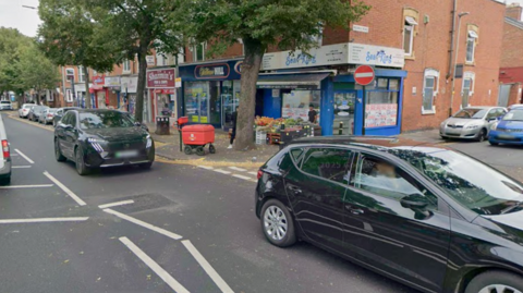 Shops and homes in Melton Road and Flax Road in Rushey Mead, with many cars driving on the road