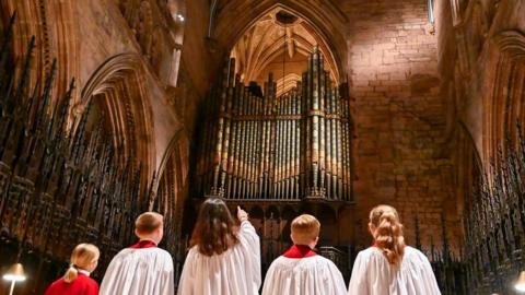 Five people from behind, looking up at the organ pipes which are black and gold. They are wearing white robes with red collars and the girl in the middle, who has brown long hair, is pointing at the pipes. The church surrounds them with stone archways on either side.