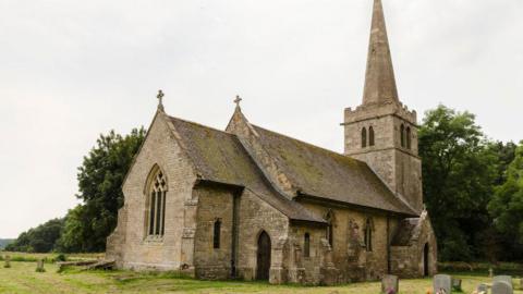 A church with a spire. It has a churchyard, and there are trees on the perimeter.