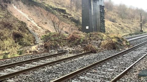 A landslip across the railway line with a bridge in the background
