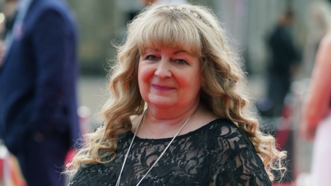 Comedian Janey Godley with long blonde hair and a hair band smiling at the camera. She is wearing a black lace top and a long silver necklace.