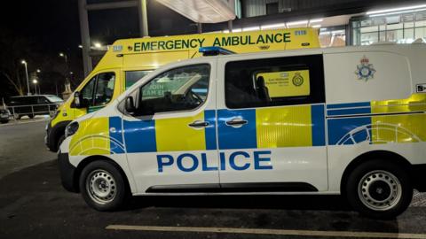 A white police van with yellow and blue cheques and the word "Police" written on the side in blue letters. Behind it is a yellow ambulance with the words "Emergency Ambulance" on the side. They are parked outside a hospital at night. 