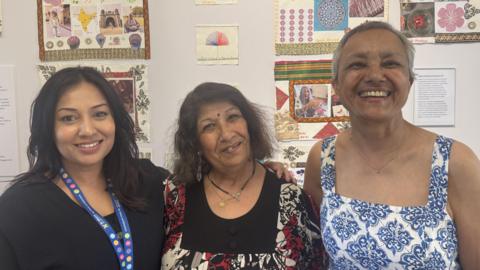 Three people stand close together indoors in front of a wall decorated with colourful artwork.