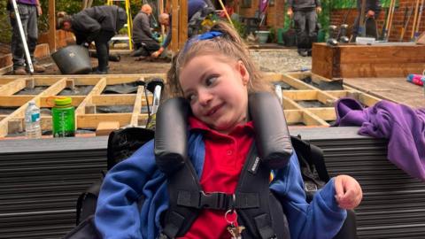 A girl smiling. She is sitting in a wheelchair and has a blue jacket with a red top on. She has brown hair and two bows in her hair. Behind her are workmen building a new garden. 
