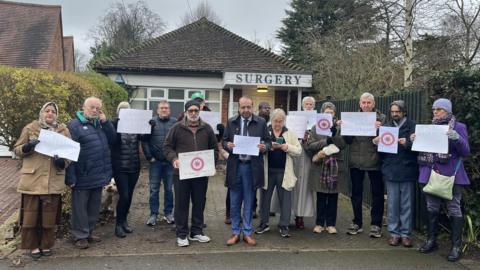 A row of people stood on the pavement holding protest signs in front of a building with a sign that reads surgery.