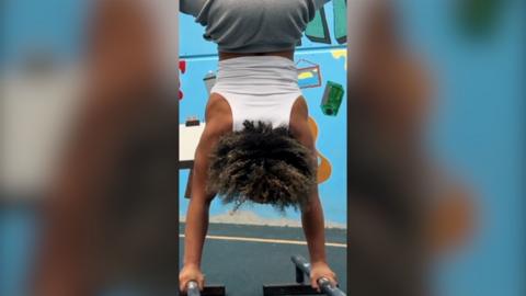 A woman wearing a white vest top and grey jogging bottoms does a handstand using two metal bars.