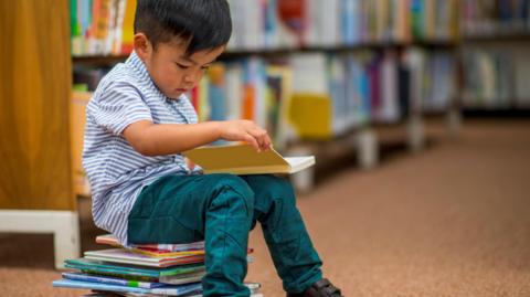 Boy with black straight hair sitting on a pile of books reading.