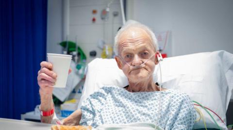 A man in his late-seventies sits up in a hospital bed and raises a paper cup up to the camera. 