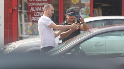 A man with a black hat, blue sunglasses and wearing a short-sleeved shirt stands behind a car while looking at a phone. A man next to him in a white shirt holds out his hand to the phone. 