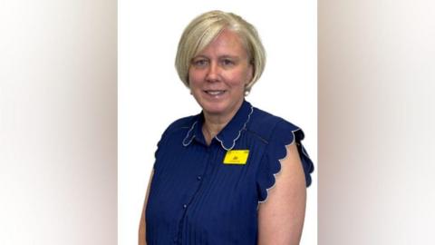 A woman with a short white blonde bob hairstyle is wearing a navy blue blouse and pearl earrings. She has a yellow name badge on that reads "Jo".
