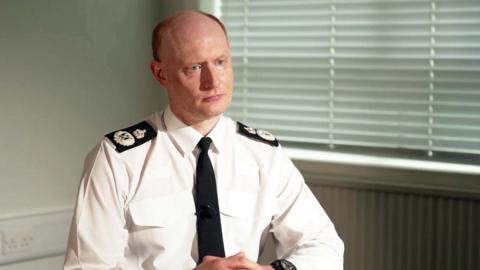 Chief Constable Alexis Boon wearing a white shirt with black epaulettes and a black tie sitting in front of a closed white Venetian blind