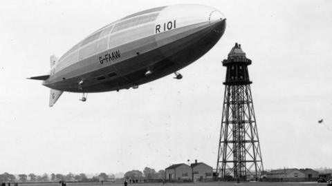 The airship R101 at its mooring mast at Cardington, Bedfordshire. It shows a large airship, in the sky, with buildings below. The image is black and white. 