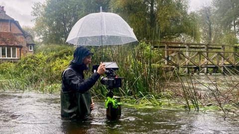 A cameraman stands waist deep in a river under an umbrella