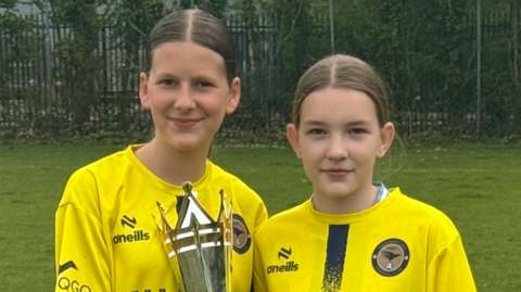 Nicole and Lexi stand side-by-side on the pitch in their yellow football kit holding a large silver trophy between them