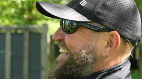 A picture of Jonny Yaxley laughing. He has three looped earrings in his left ear and is wearing sunglasses. He is wearing a black cap.