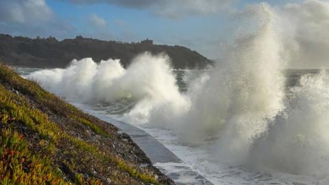 Huge waves crash on to the beach in Falmouth, Cornwall, during Storm Ingrid, with masses of very high foamy water approaching the land to the left and a headland behind