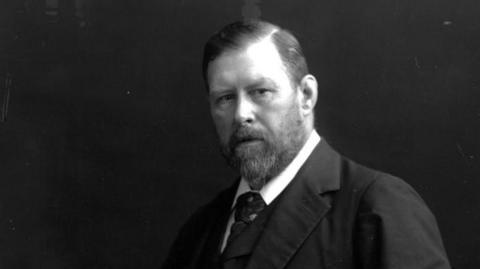 A black and white photo of author Bram Stoker from 1890. He wears a dark coloured suit, white shift and dark coloured tie. He has a short beard and a side-parted hairstyle. 