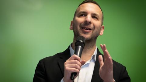 Zack Polanski holding a microphone in front of a green background. He has an open-necked shirt.