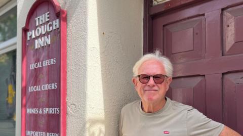 An man with grey hair, wearing a beige levi's t shirt and sunglasses. He stands in the doorway of a pub, next to a red sign with the "plough inn" and types of alcohol on it.