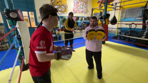 Two young students practice with sparring gloves and pads in kickboxing ring. Sam Byford-Winter, co-founder of Nam Yang, stands outside the ring leaning on the ropes and watches them.
