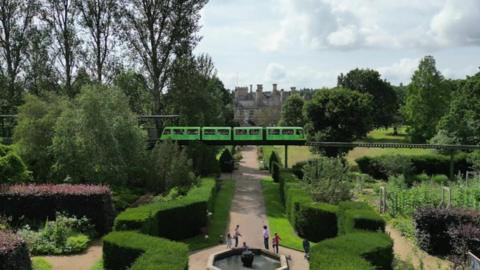 Stately home gardens with a fountain and well pruned bushes. A monorail line is above with a green train travelling along it. 