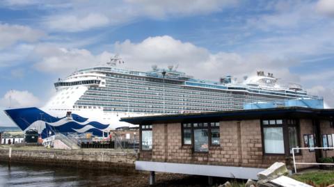 A large cruise ship is berthed at Invergordon. The ship has several floors and is blue and white in colour. There is an office in the foreground.