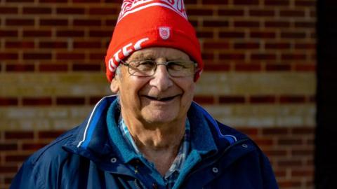 A man wearing a blue anorak, a red hat and a pair of glasses, smiling at the camera in front of a brick wall