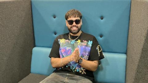 A mean wearing dark sunglasses inside, a colouful t shirt and chains around his neck sits in a huge blue and grey chair