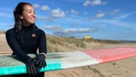 A woman with dark hair, wearing a wet suit is sat smiling and holding a green, white and red surf board