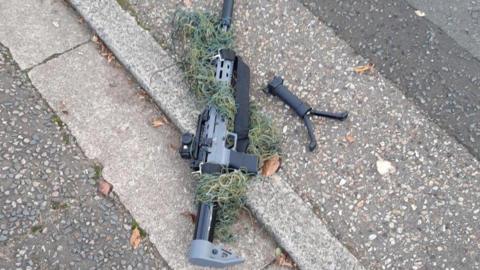 A grey and black air rifle placed on a concrete road. The rifle handle is detached from the gun, loose on the ground.