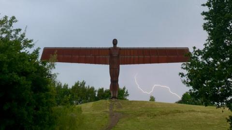The Angel of the North: a rust-coloured statue of an angel with extended wings. It stands on a hill, with greenery on either side. Lightning can be seen, seemingly rising from a bush beneath the right wing of the sculpture.