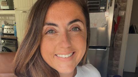 Rebecca Allerton looks up at the camera while taking a selfie. She is stood in a kitchen and behind her is a large silver fridge with a picture on it drawn by a child.