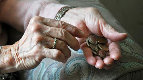 A female pensioner's hands. She is wearing a ring on her right ring finger and a watch on her left arm. In her left hand there are £1 and 50p coins. Her hand is resting on a sofa.