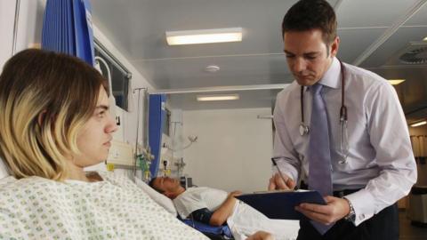 A female patient in a white gown with a green pattern on it is lying in bed. She has dark hair with blonde tips. A doctor wearing a white shirt and a purple tie is standing next to her, writing on a clipboard. He has brown hair and has a stethoscope round his neck. In the background is a hospital ward, with a man lying on a hospital bed