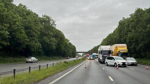 A northbound and southbound carriageway, both being three lanes, are visible in the image, with the right hand carriageway featuring cones across the road stopping traffic. You can see some cars and lorries at the front of the traffic queue. 