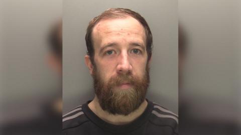 Police custody image of Lee Merrison he has short brown hair and a beard and is wearing a dark shirt with grey stripes on the shoulders