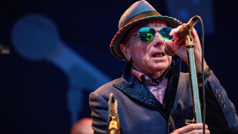 Sir Van is singing onstage. He's standing in front of a mic on a stand. He's wearing sunglasses, a hat and a blue jacket with a pink shirt.