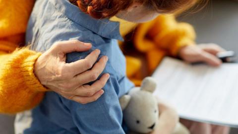 A woman wearing an orange jumper sitting with her arm around a young girl. The child is wearing a blue shirt, has braids in her hair and is hugging a cream teddy bear while they both look at a piece of paper in the woman's hand 